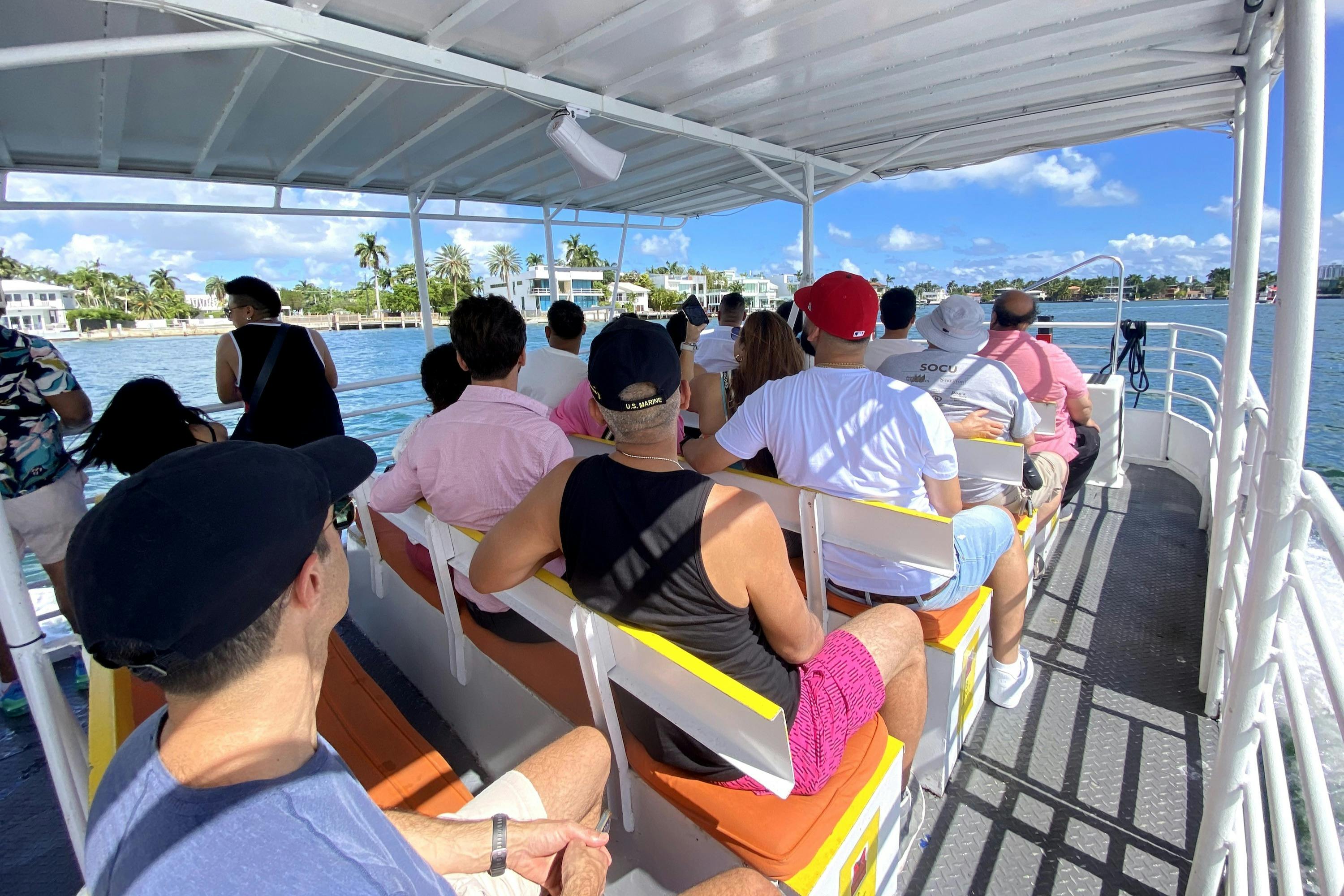 Folk sidder på en overdækket båd, der vender fremad. Baggrunden viser vand, palmer og hvide bygninger under en blå himmel.