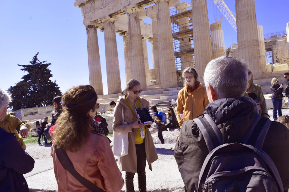 A group of people gathered near the Parthenon in Athens, some listening to a guide holding a tablet. Scaffolding visible.