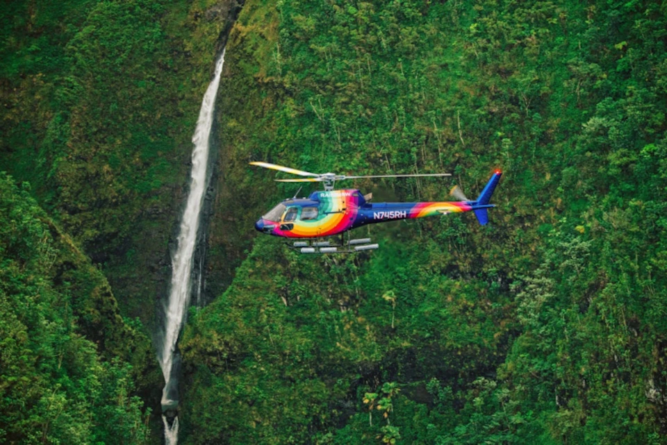 A rainbow-colored helicopter flies near a waterfall against a backdrop of dense greenery.