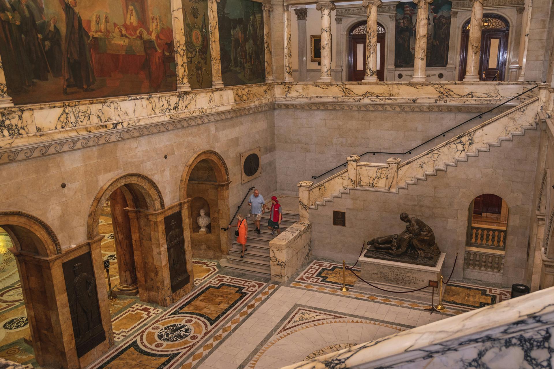 People walking in a marble-floored museum with statues, archways, and large paintings on the walls.