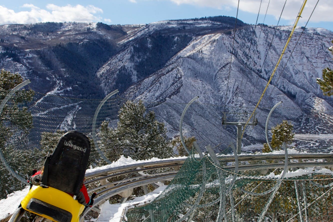Un trineu en una pista de muntanya russa en un paisatge de muntanya nevada amb arbres i una xarxa de seguretat.