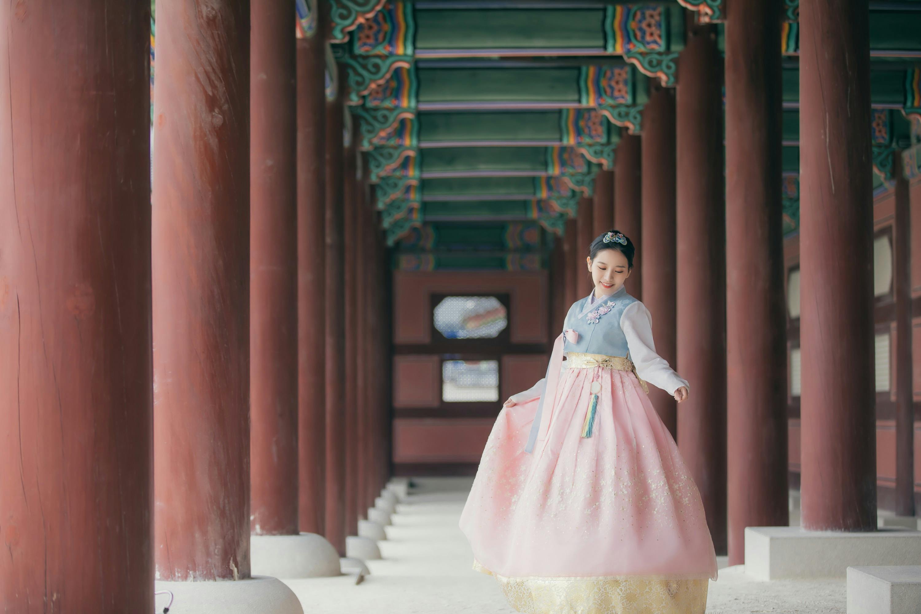 A woman wearing a traditional hanbok stands gracefully in a corridor of a historic building with colorful wooden beams.