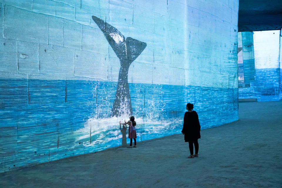 A child and an adult observe a large wall projection of a whale's tail splashing in the ocean.