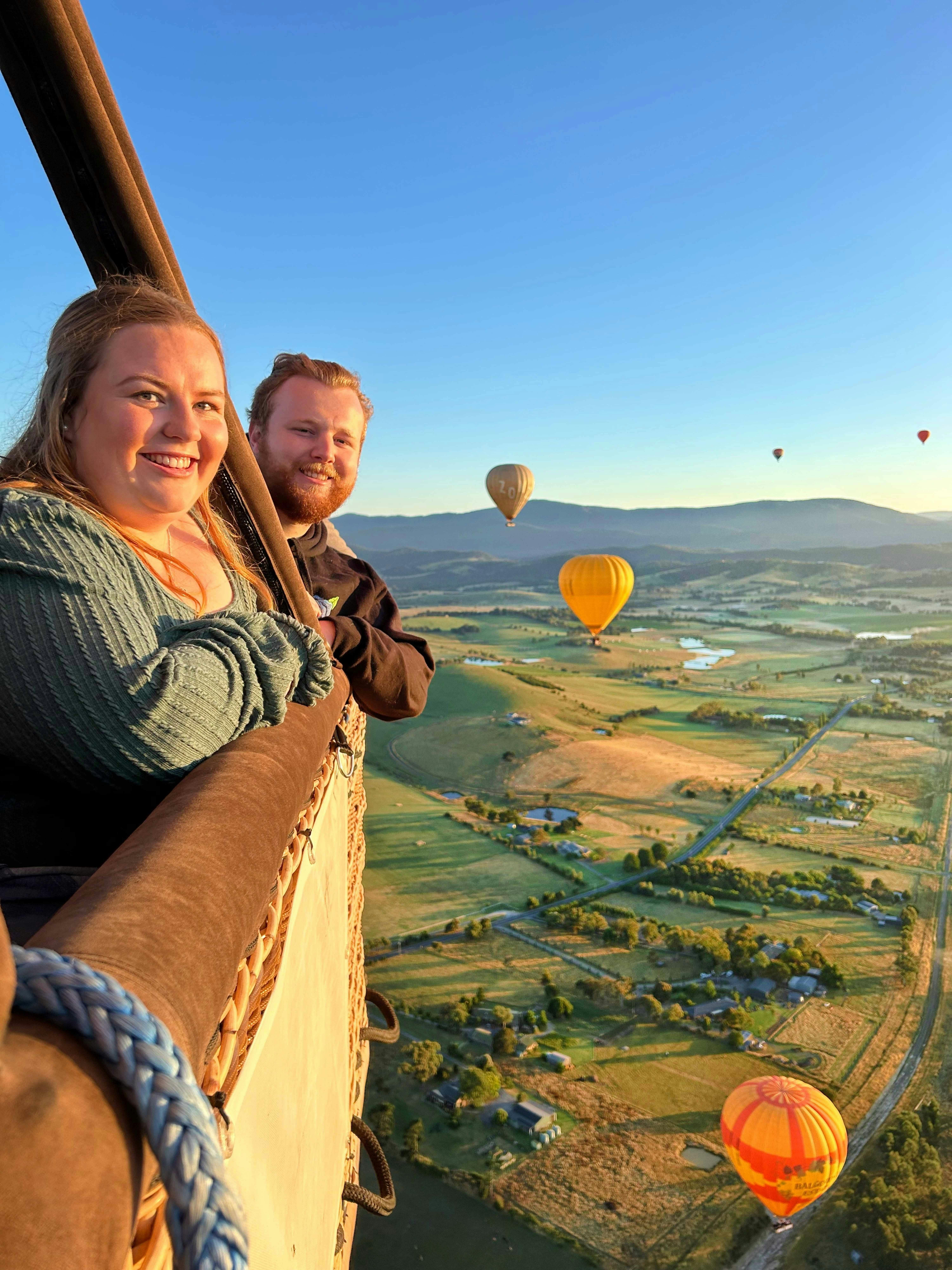 Two people smile while riding in a hot air balloon, overlooking a landscape with multiple other hot air balloons in the distance.
