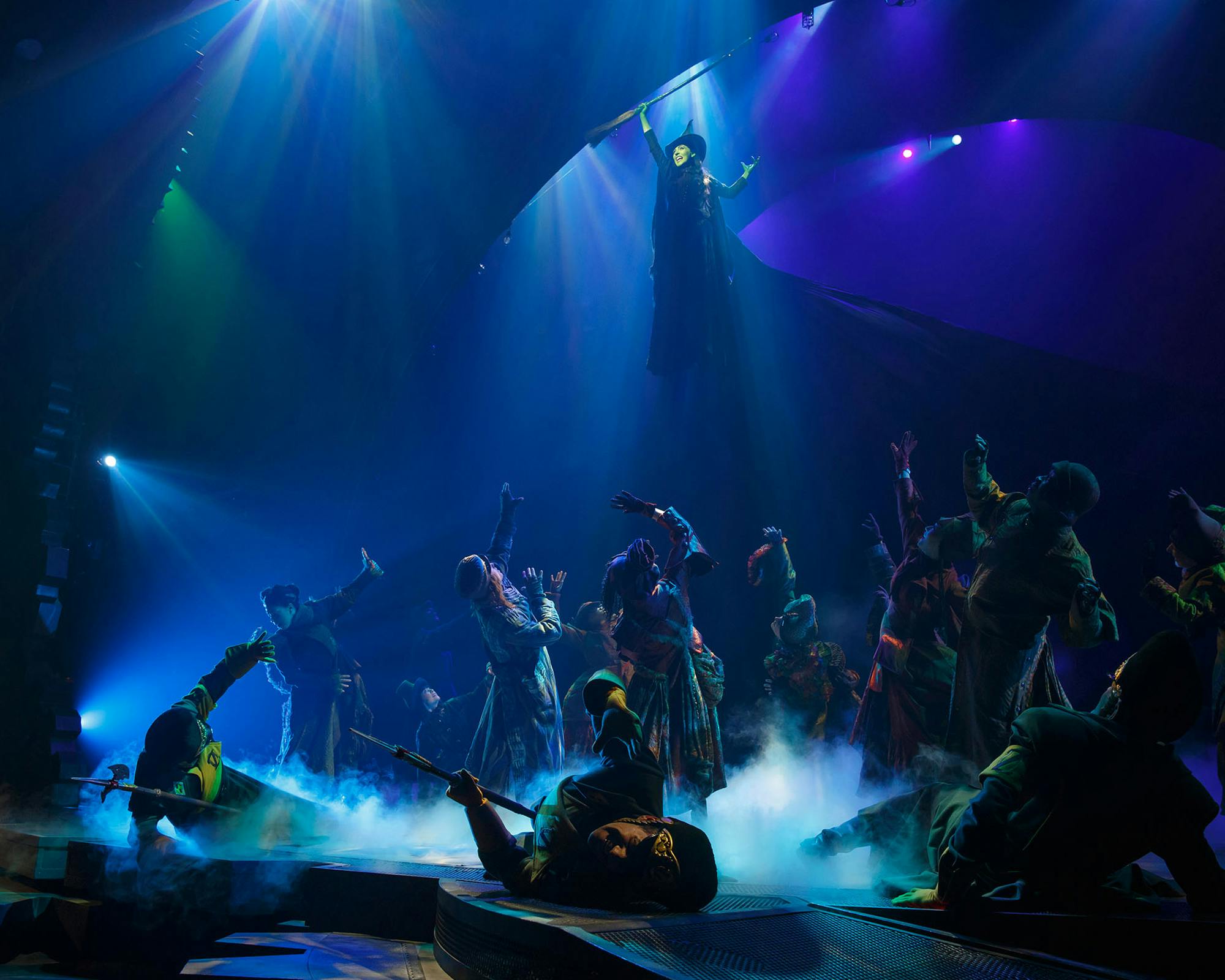 舞台上的表演者，有雾气效果和五颜六色的灯光；一个人被悬挂在组上方，大幅伸展手臂。