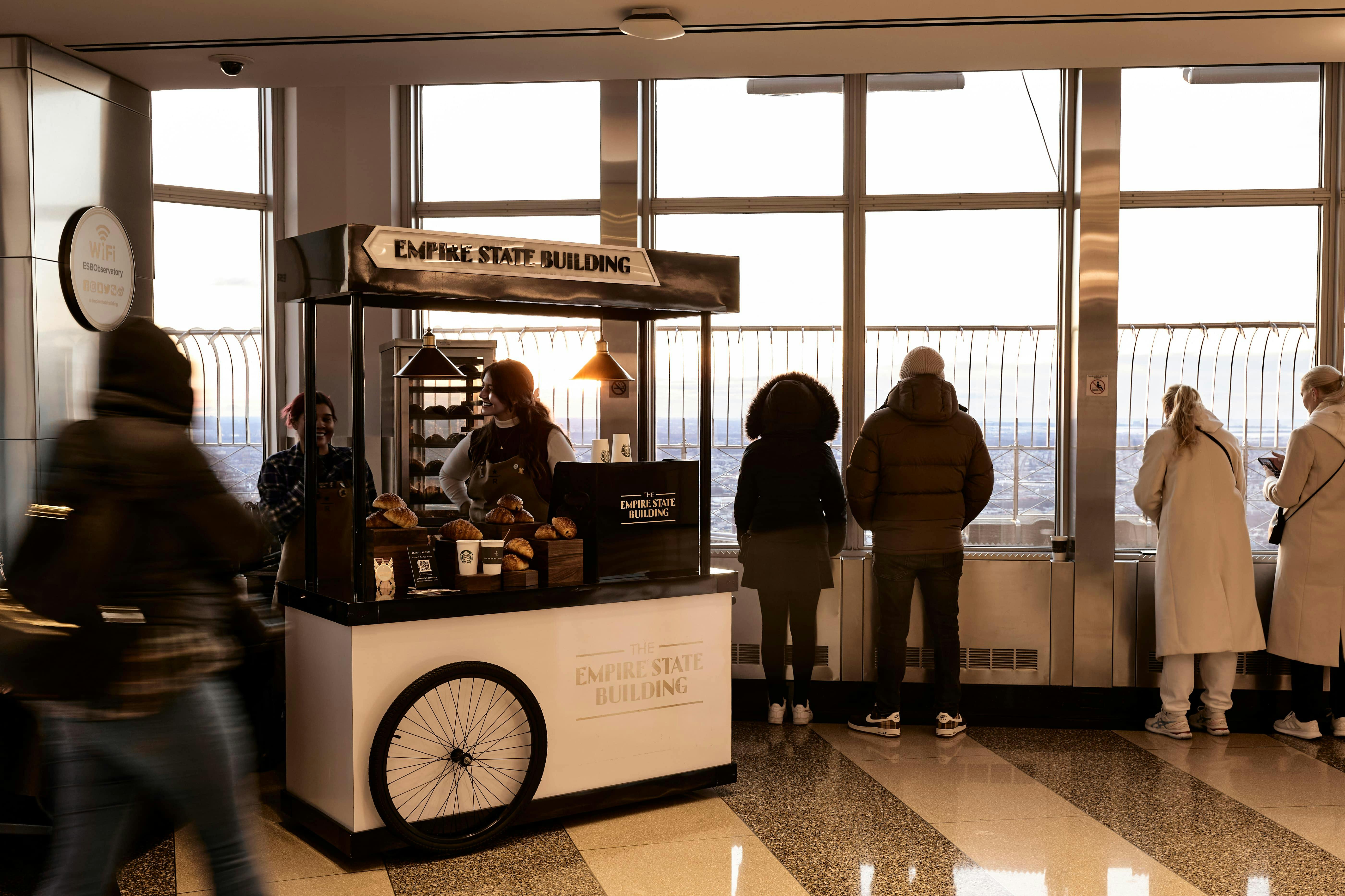 People stand by a window near a snack cart labeled "Empire State Building," featuring pastries and coffee.