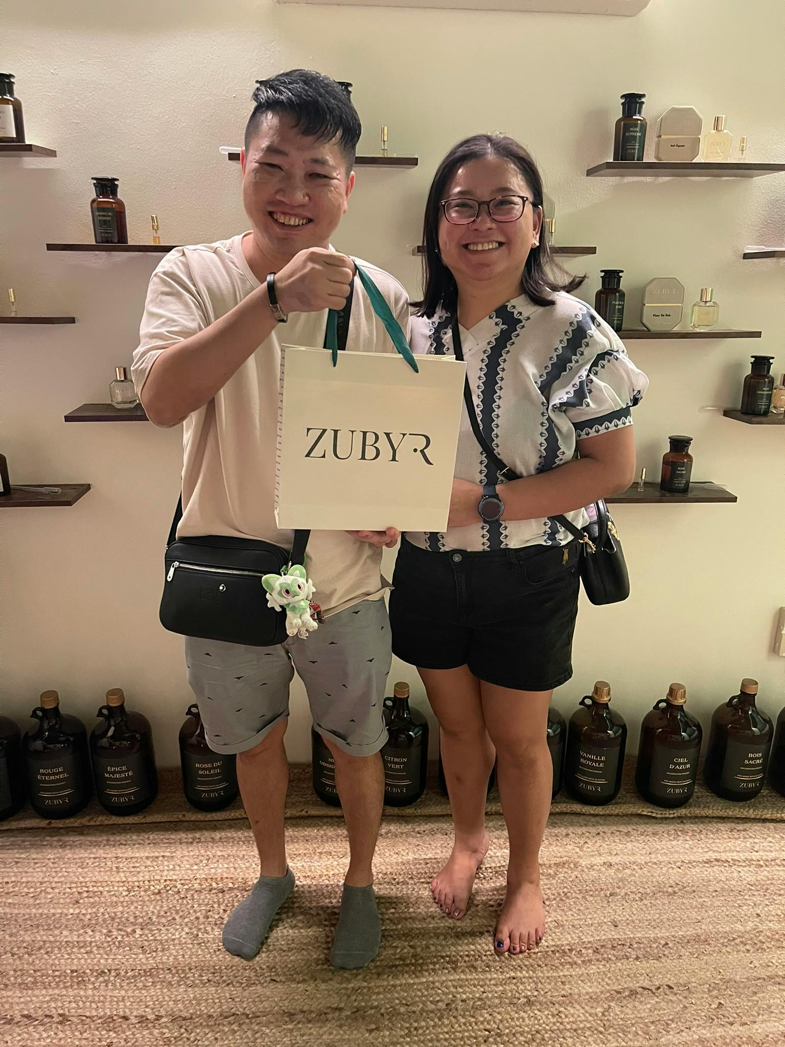 Two people smiling and posing with a branded shopping bag in front of shelves displaying bottles and products.