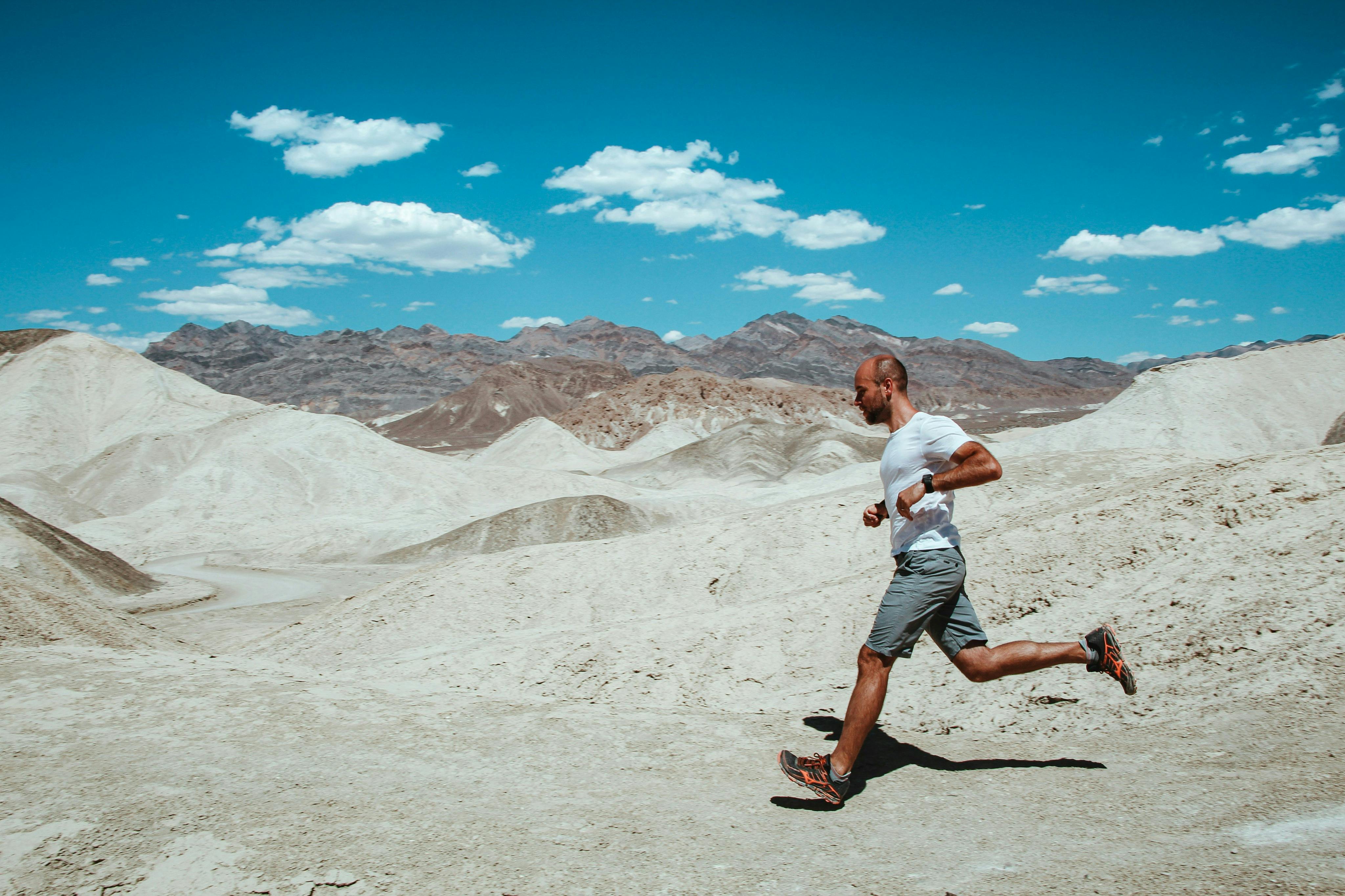A man jogging on a rocky desert terrain under a clear blue sky with scattered clouds and mountains in the background.