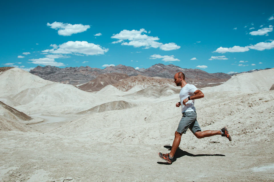 A man jogging on a rocky desert terrain under a clear blue sky with scattered clouds and mountains in the background.