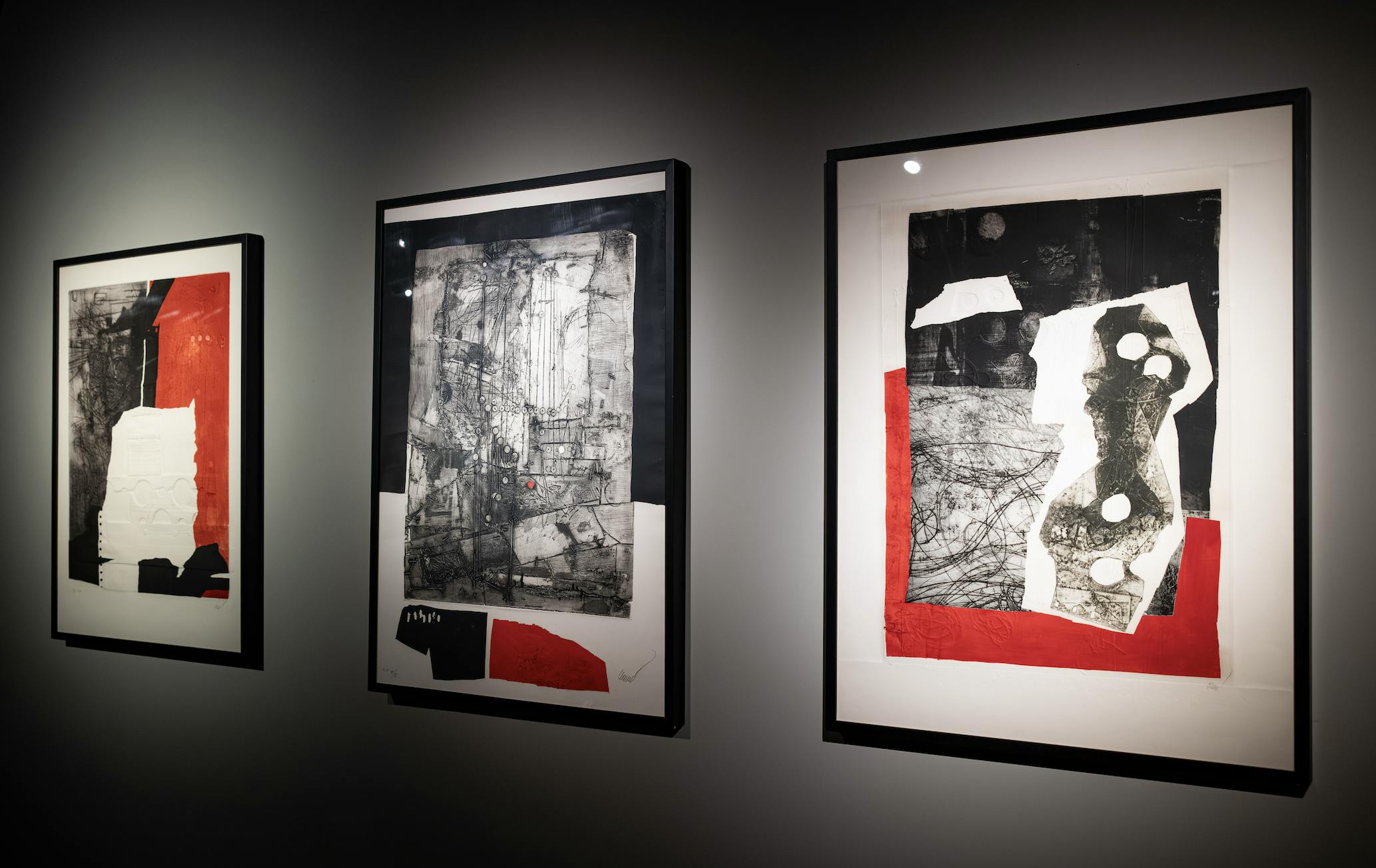 Drie ingelijste abstracte kunstwerken op een donkere muur, met voornamelijk zwarte, witte en rode tinten.