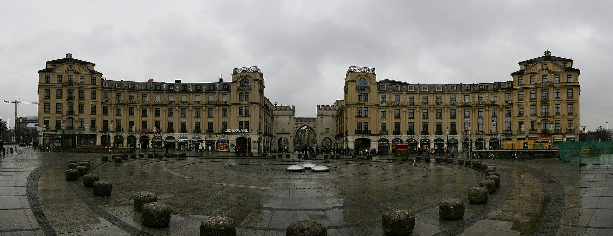Duży plac z mokrym, okrągłym kamiennym brukiem, otoczony beżowymi zabytkowymi budynkami i łukami pod zachmurzonym niebem.