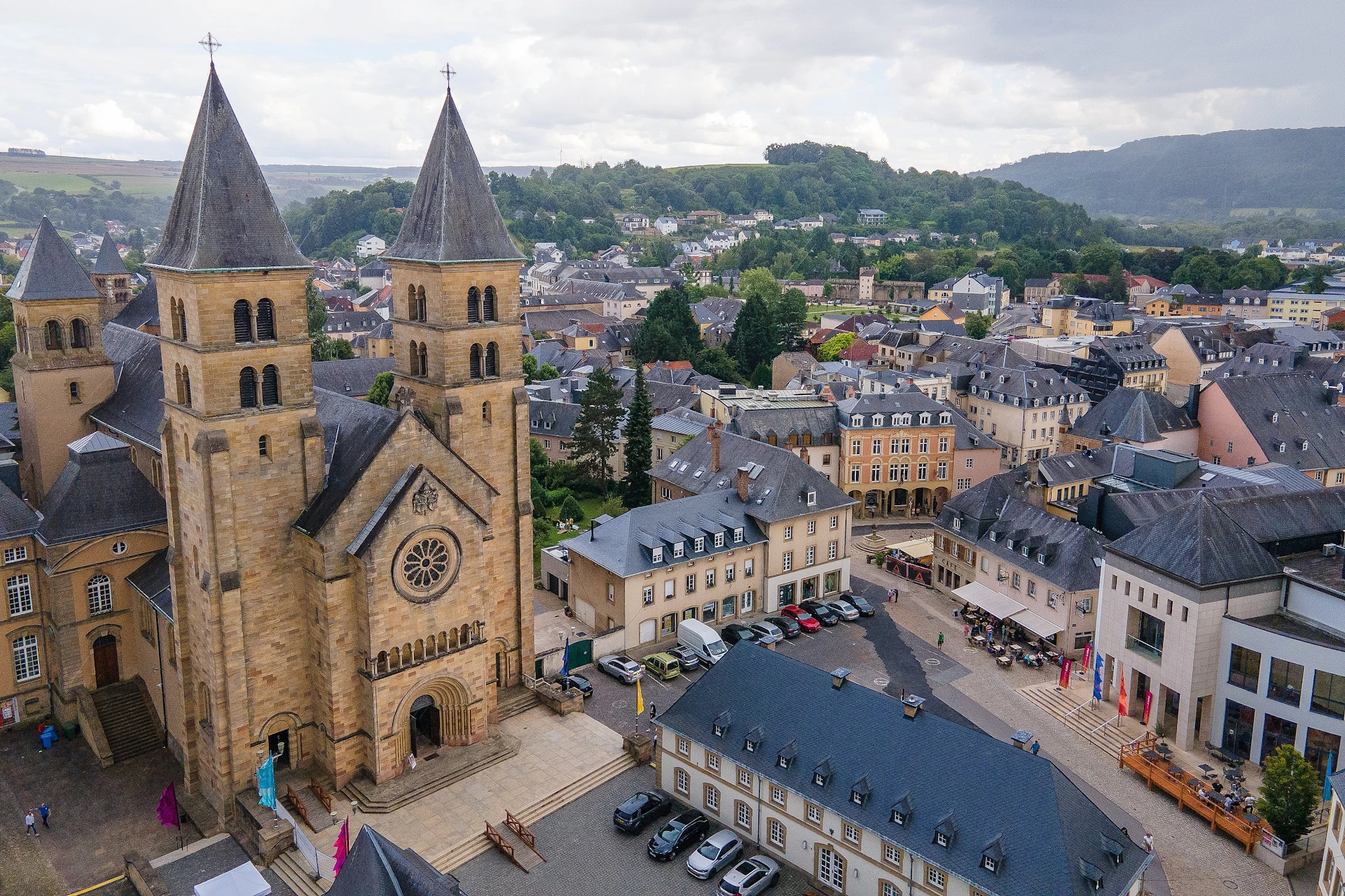 Aerial view of a European town with a church featuring twin spires, surrounded by buildings and greenery in the background.