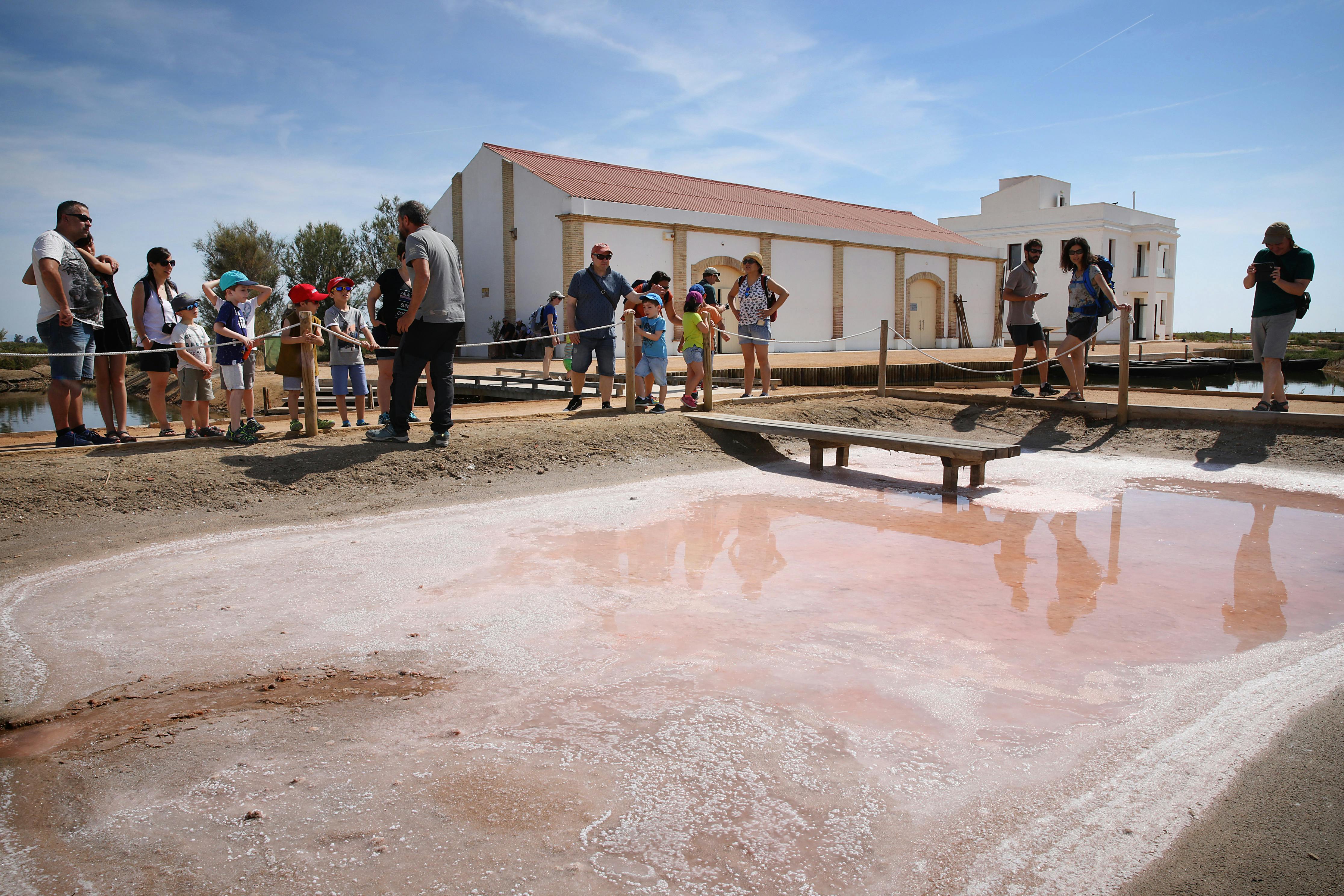 Οι άνθρωποι εξερευνούν ένα αλμυρό διαμέρισμα με ροζ επιφάνεια, ξύλινες γέφυρες και ένα κτίριο με κόκκινη στέγη στο παρασκήνιο.