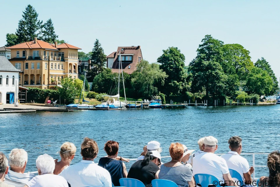 People seated on a boat facing a scenic view with houses, sailboats, trees, and clear blue water under a bright sky.