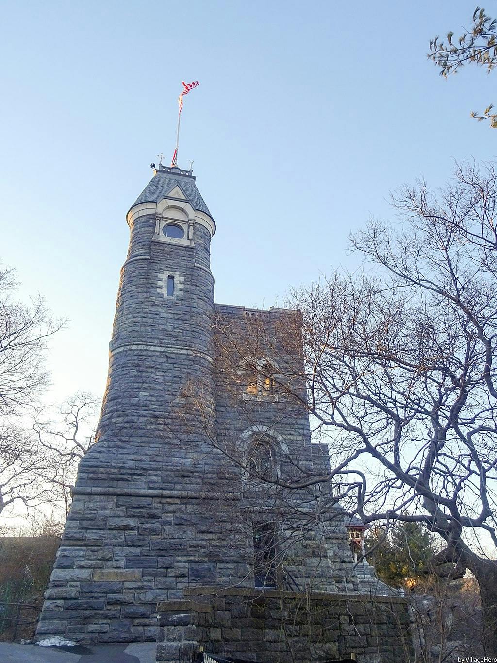 Stone tower with a narrow turret, topped by a flag, surrounded by bare trees under a clear sky.
