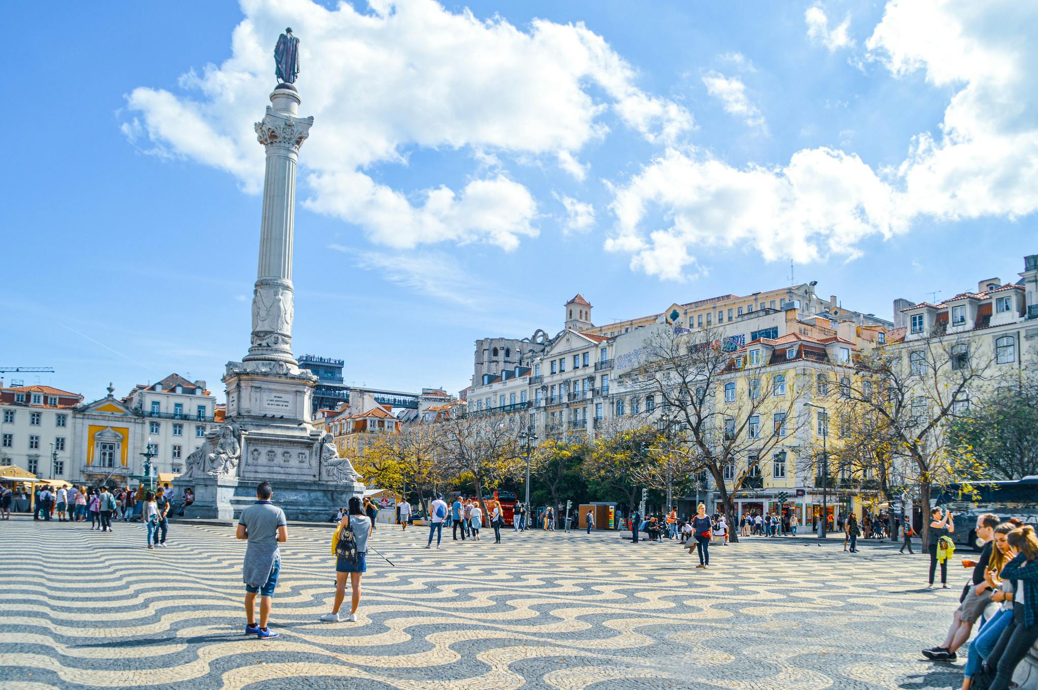 Náměstí s vysokým bílým pomníkem, vzorovanou dlažbou, okolními budovami a lidmi, kteří chodí a fotí.