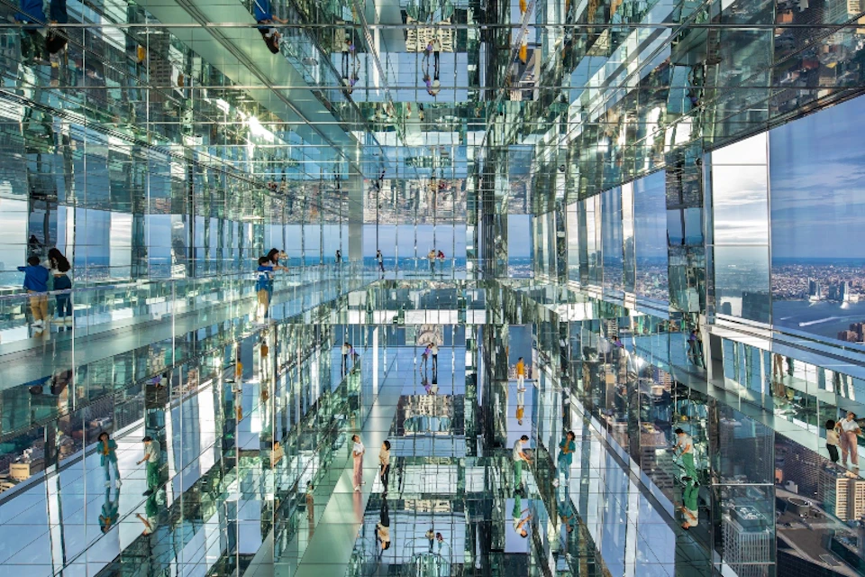 People walk and take photos in a room with mirrored walls and floors, creating multiple reflections. Cityscape is visible through windows.