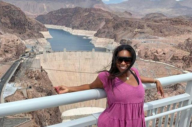 A person in a pink outfit stands on a railing with a scenic view of a dam and rocky landscape in the background.