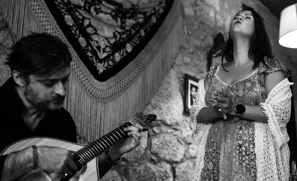 A seated man plays a guitar while a standing woman sings with eyes closed and hands clasped in a stone-walled room.