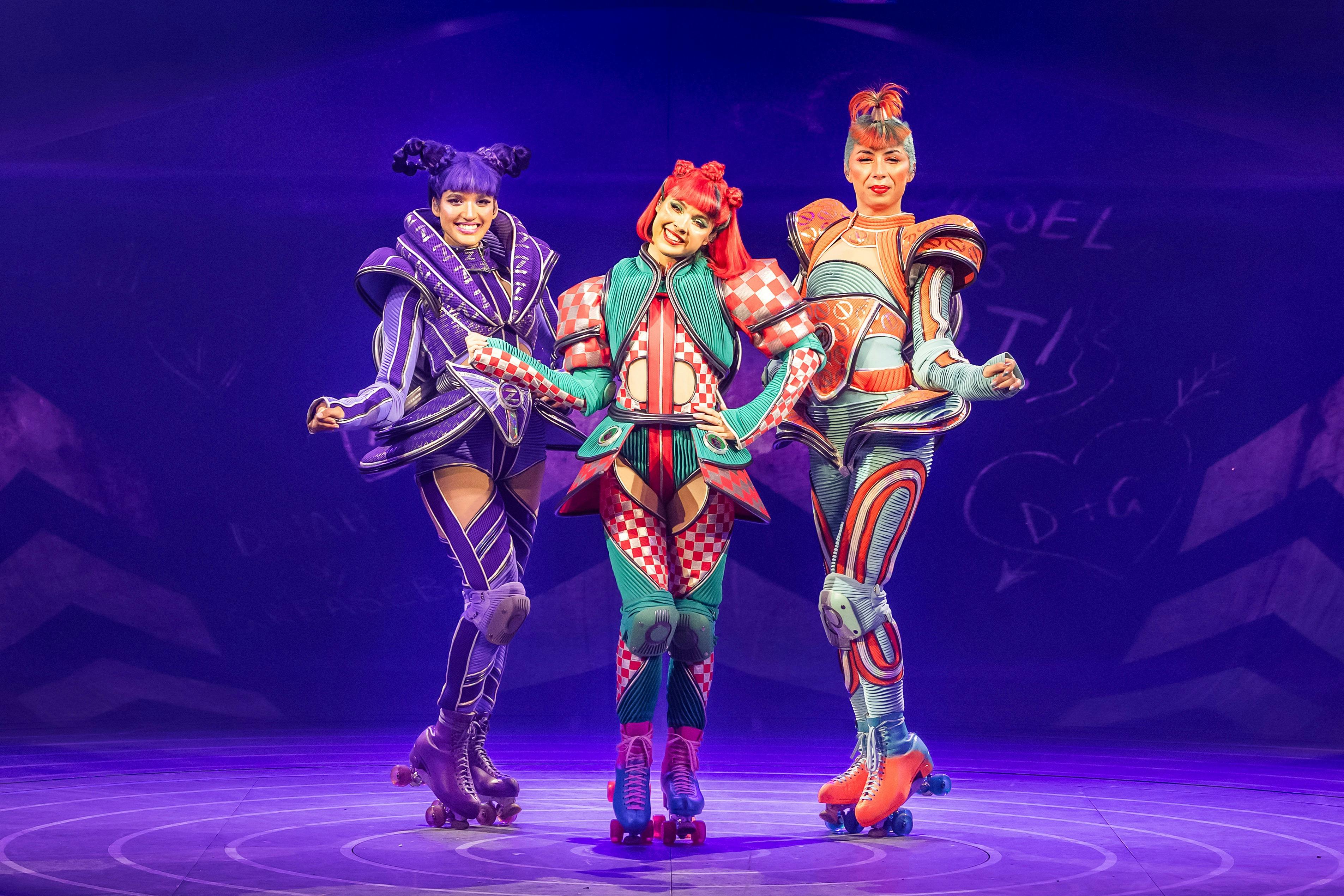 三位穿着充满活力、充满未来感的服装和溜冰鞋的表演者在涂鸦的背景下在舞台上微笑。