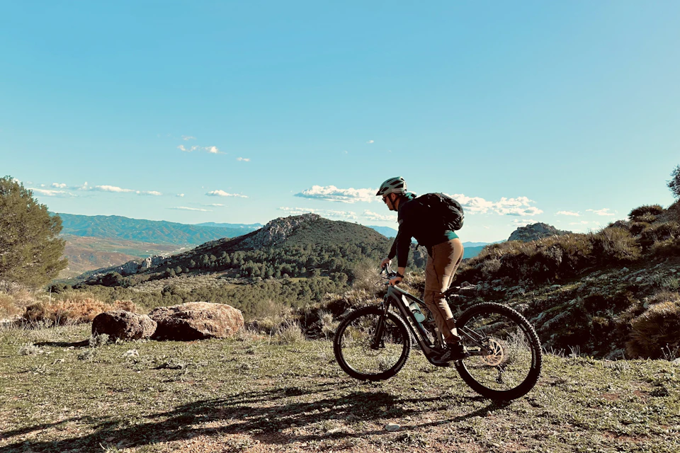 A person riding a mountain bike on a grassy trail with hills and a clear blue sky in the background.