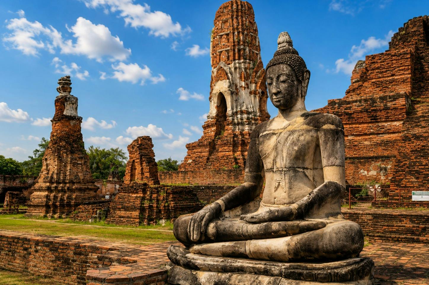 A large stone Buddha statue in a seated pose at an ancient brick temple under a blue sky with scattered clouds.