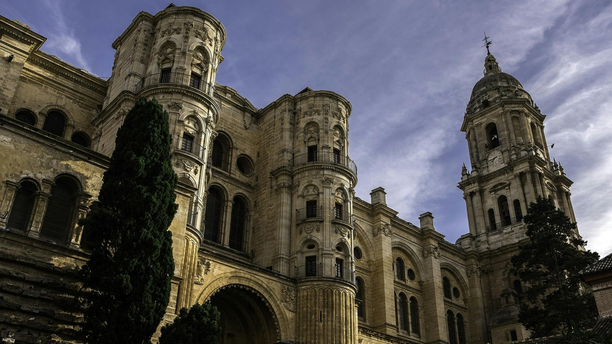 Historical stone building with arched windows, detailed carvings, and a tall clock tower under a partly cloudy sky, flanked by trees.