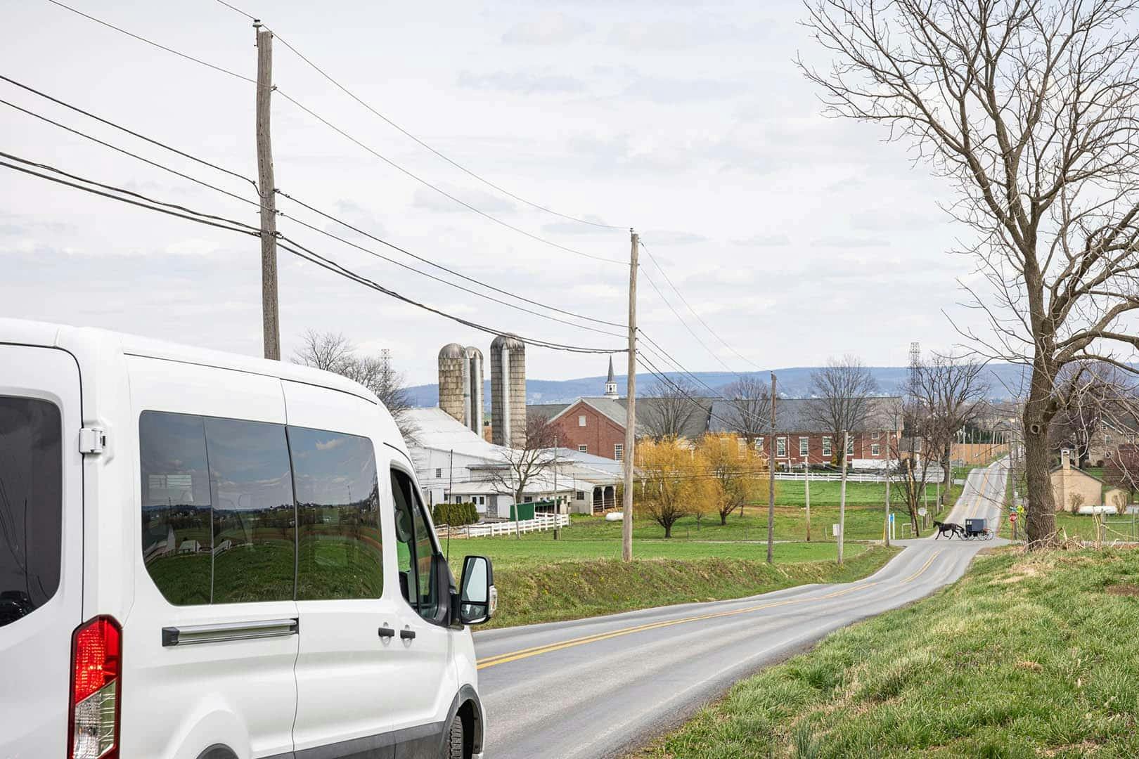 A white van drives on a rural road towards a farm with silos, barns, and homes surrounded by fields and trees.