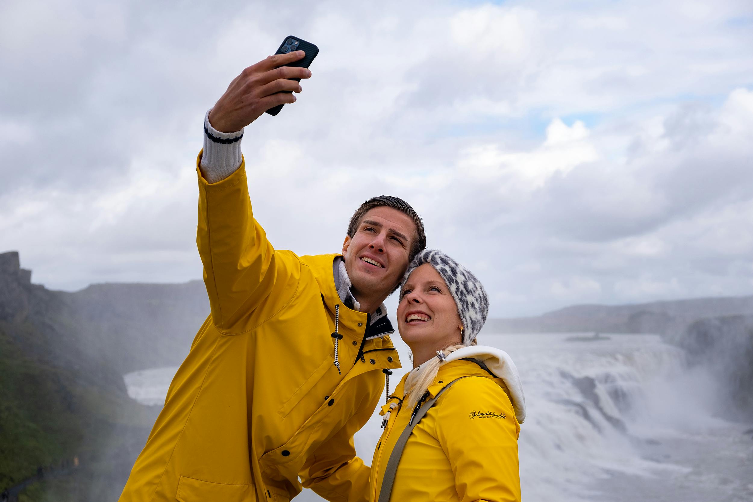 Two people in yellow jackets take a selfie in front of a large waterfall under a cloudy sky.