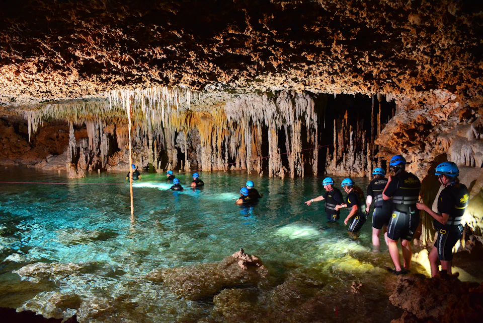 People in wetsuits and helmets exploring a water-filled cave with hanging stalactites and illuminated clear water.