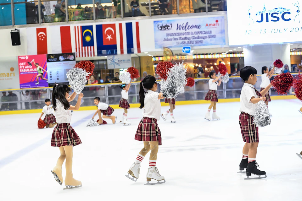 A group of young girls performing a cheerleading routine on an ice rink, holding red and silver pom-poms, with flags in the background.