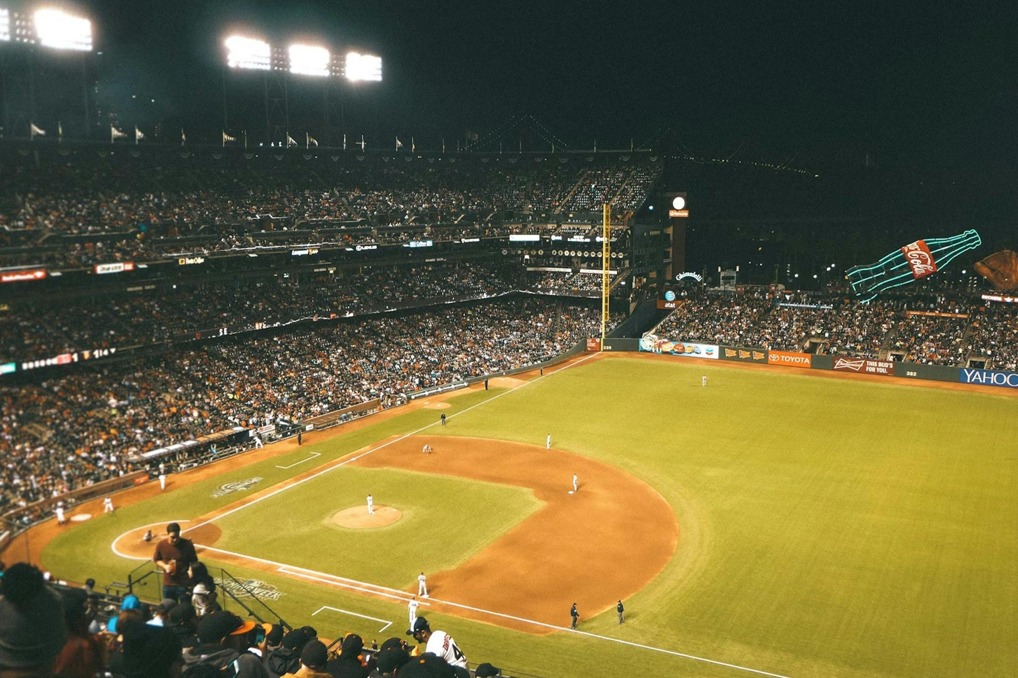 Noční baseballový zápas na přeplněném stadionu, při pohledu z tribuny, s hráči na hřišti a osvětlenými světly.