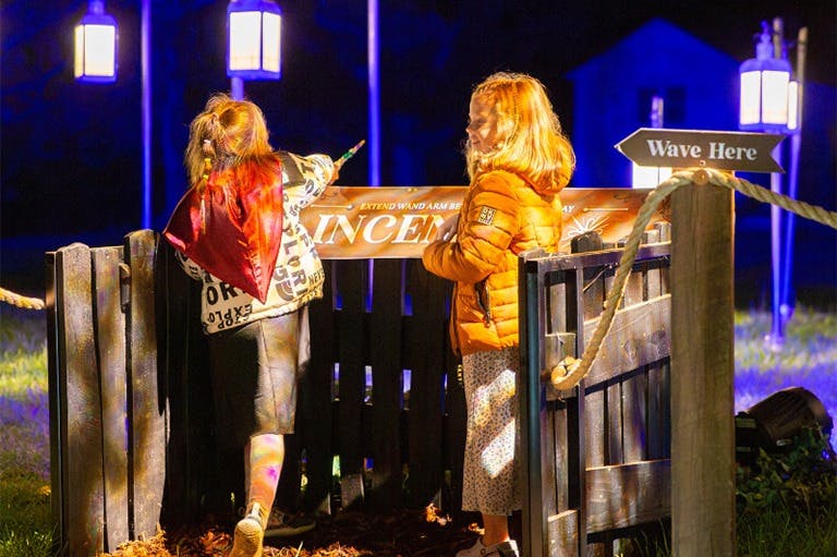 Δύο παιδιά με μπουφάν στέκονται δίπλα σε έναν ξύλινο φράχτη τη νύχτα, εν μέρει φωτισμένα από ένα φανάρι. Ο ένας δείχνει ένα ραβδί-παιχνίδι.