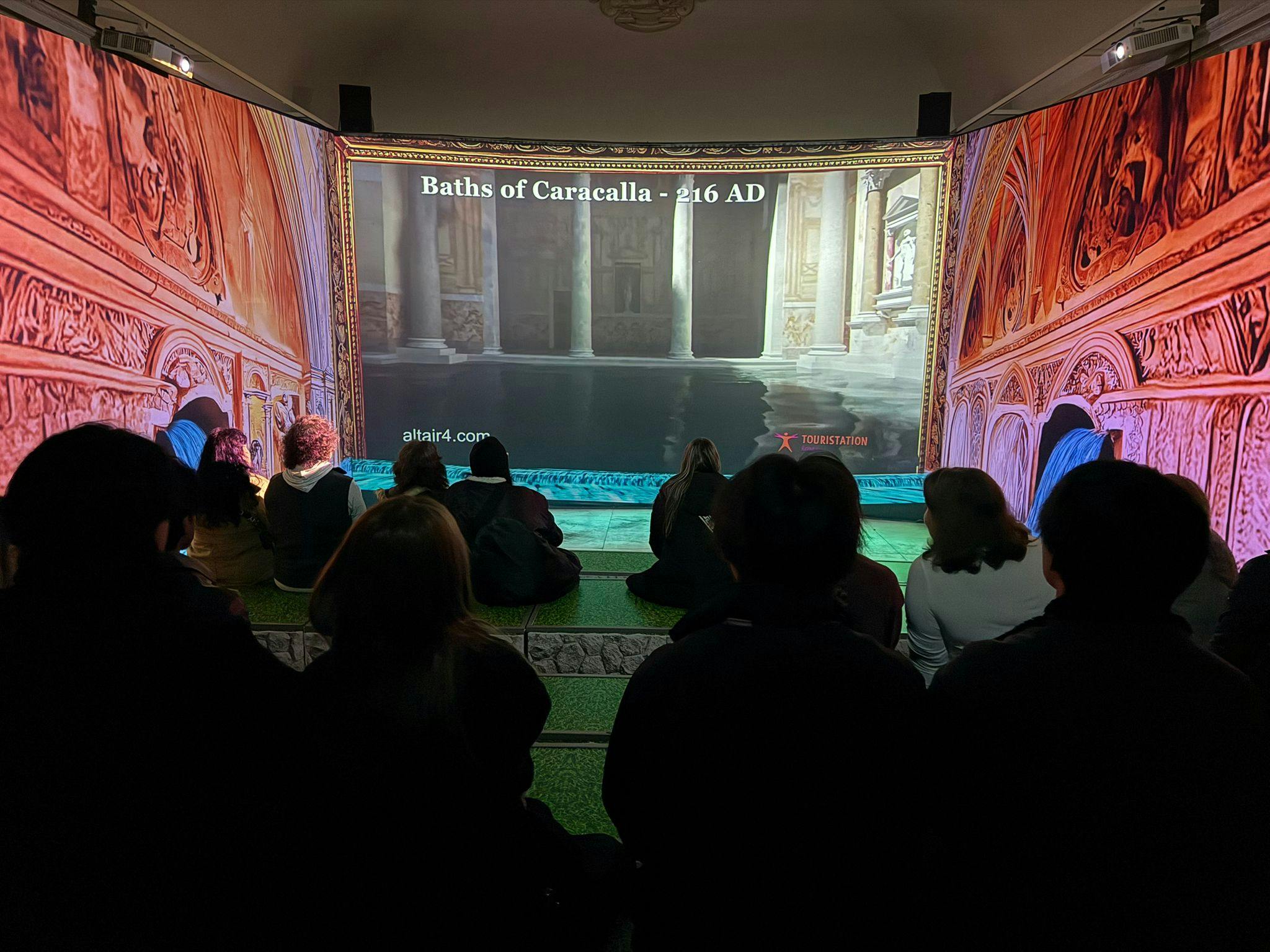 Persone sedute al chiuso che guardano una proiezione delle Terme di Caracalla, 216 d.C., visualizzate con un ambiente decorativo.