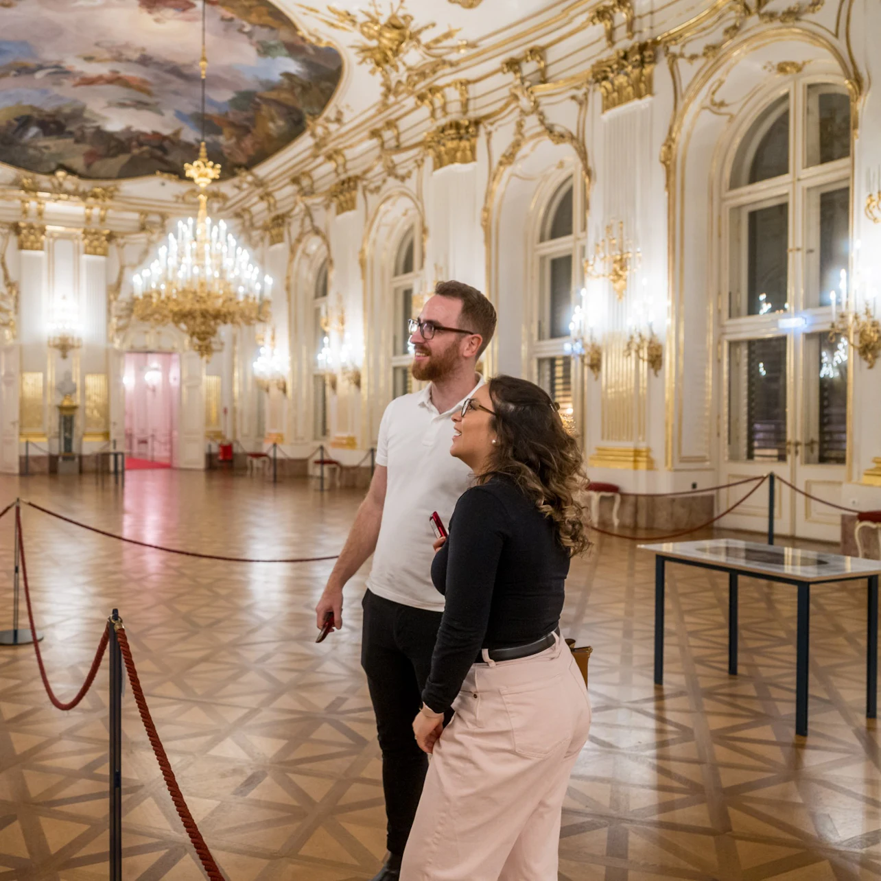 Two people admire an ornate hall with chandeliers, gilded decor, and a painted ceiling, separated by a red rope barrier.