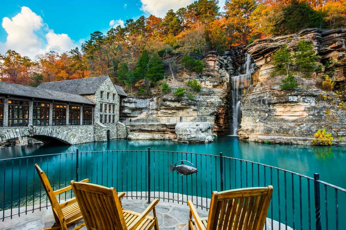 Une vue paisible sur un bâtiment en pierre au bord d'une cascade et d'une eau bleue, vue depuis une terrasse équipée de chaises en bois et d'une balustrade métallique.