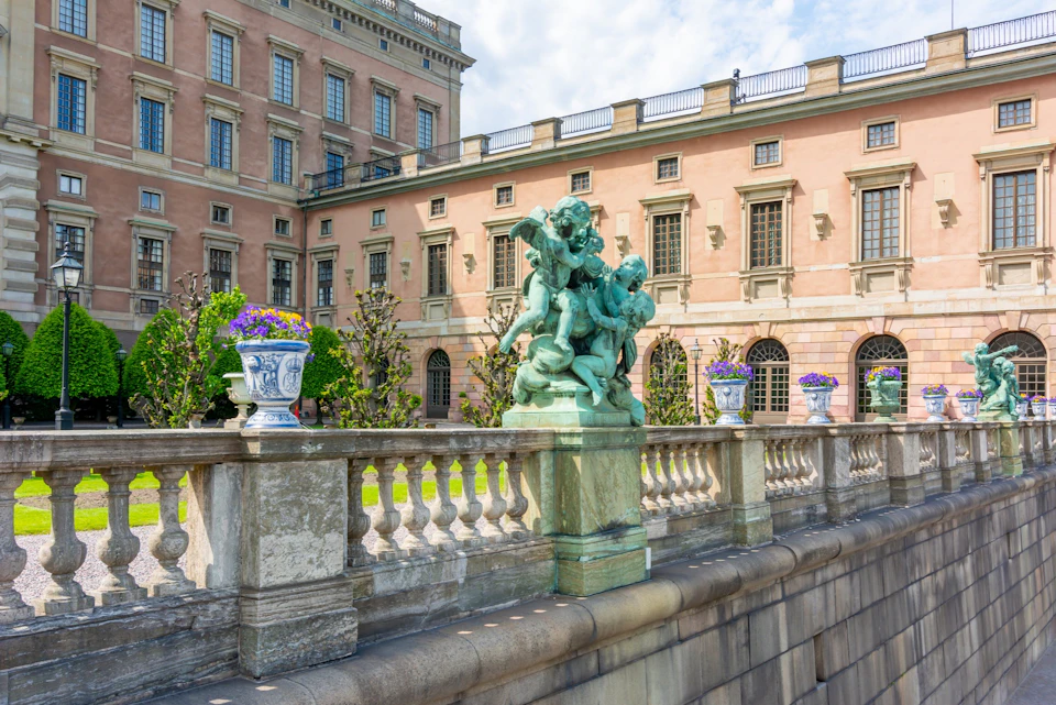 A historic building with ornate windows, a balustrade, a green statue of figures, and potted flowers in a garden setting.