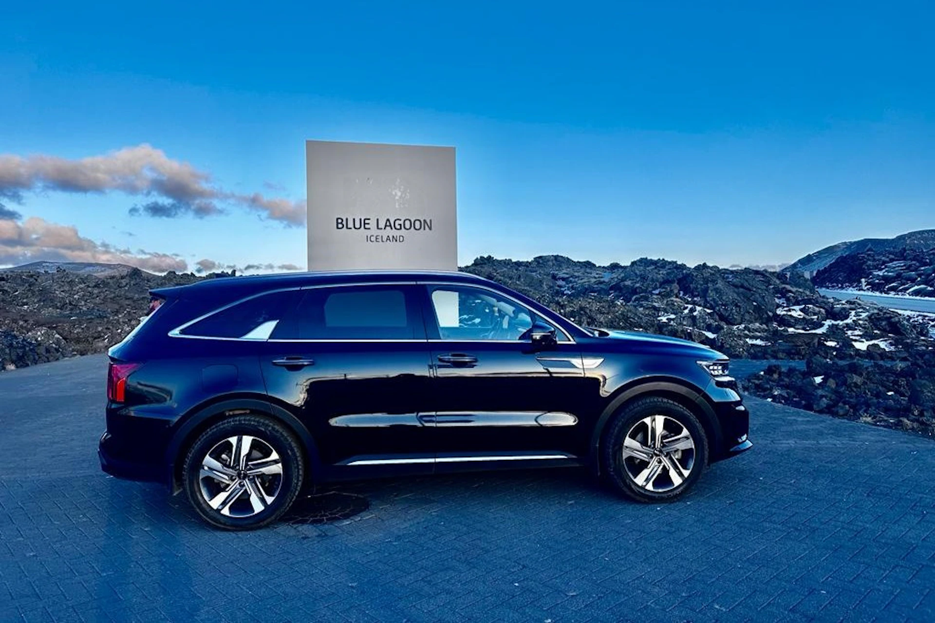 A black SUV parked in front of a sign that reads "Blue Lagoon Iceland," with a rocky landscape and a blue sky in the background.