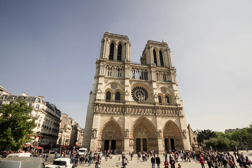 A large Gothic cathedral with twin towers and a rose window, surrounded by a plaza with people walking and gathering.