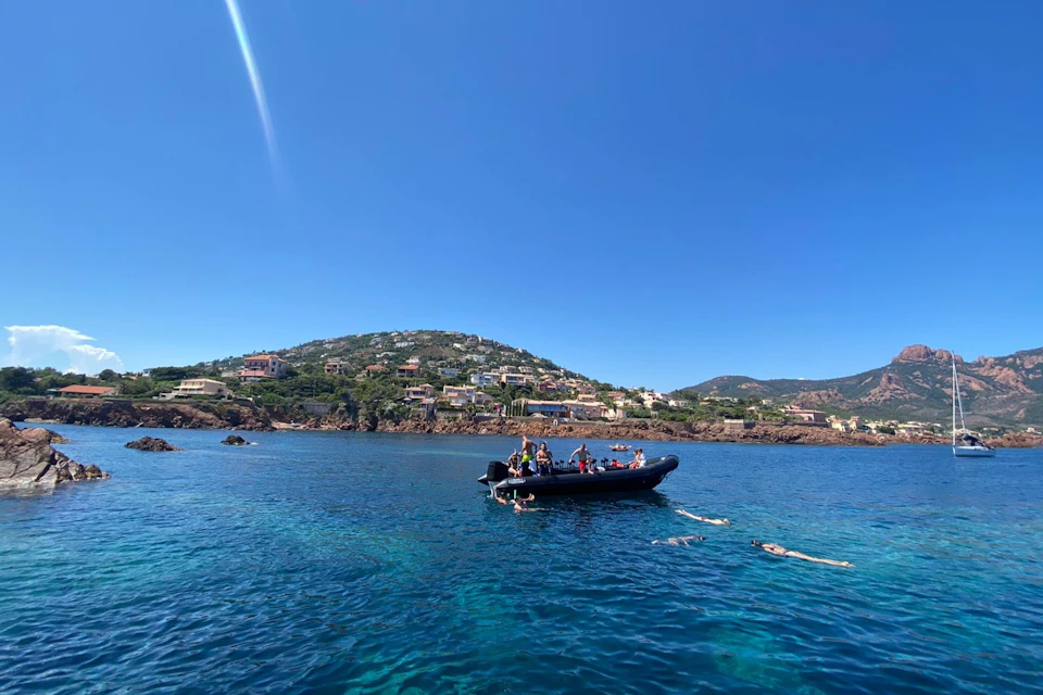 A group of people on a boat with swimmers in the water near a coastal hillside under a clear blue sky.
