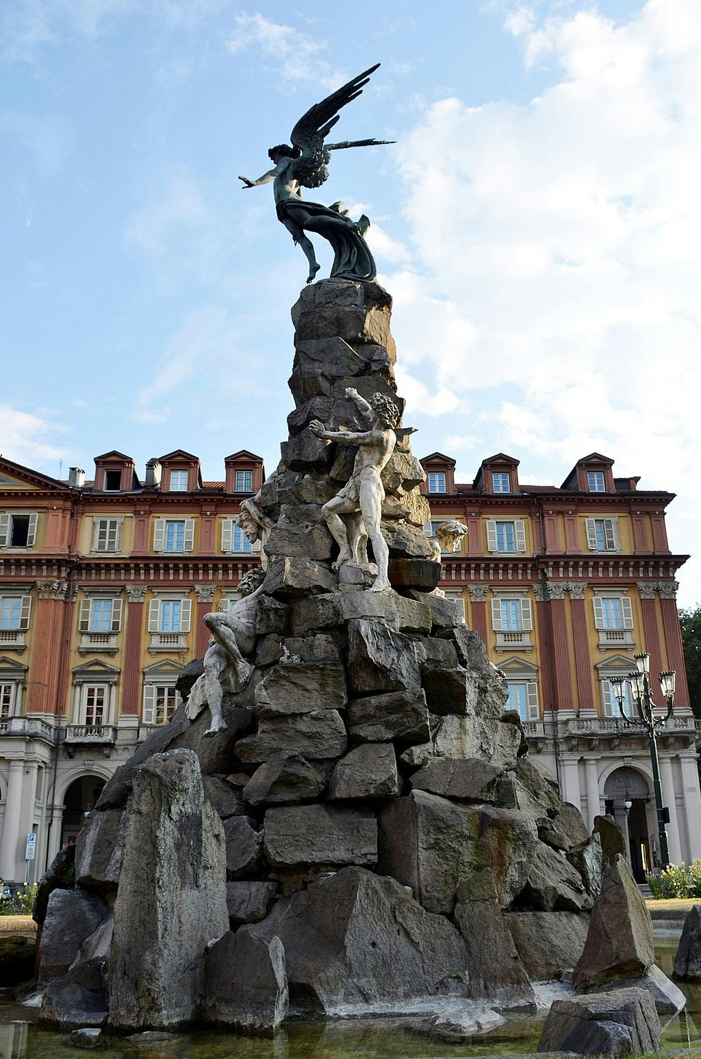Alto monumento in pietra con sculture di figure, tra cui una figura alata in cima, posto davanti a un edificio storico sotto un cielo azzurro.