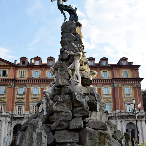 Högt stenmonument med skulpturer av figurer, inklusive en bevingad figur ovanpå, som ligger framför en historisk byggnad under en blå himmel.