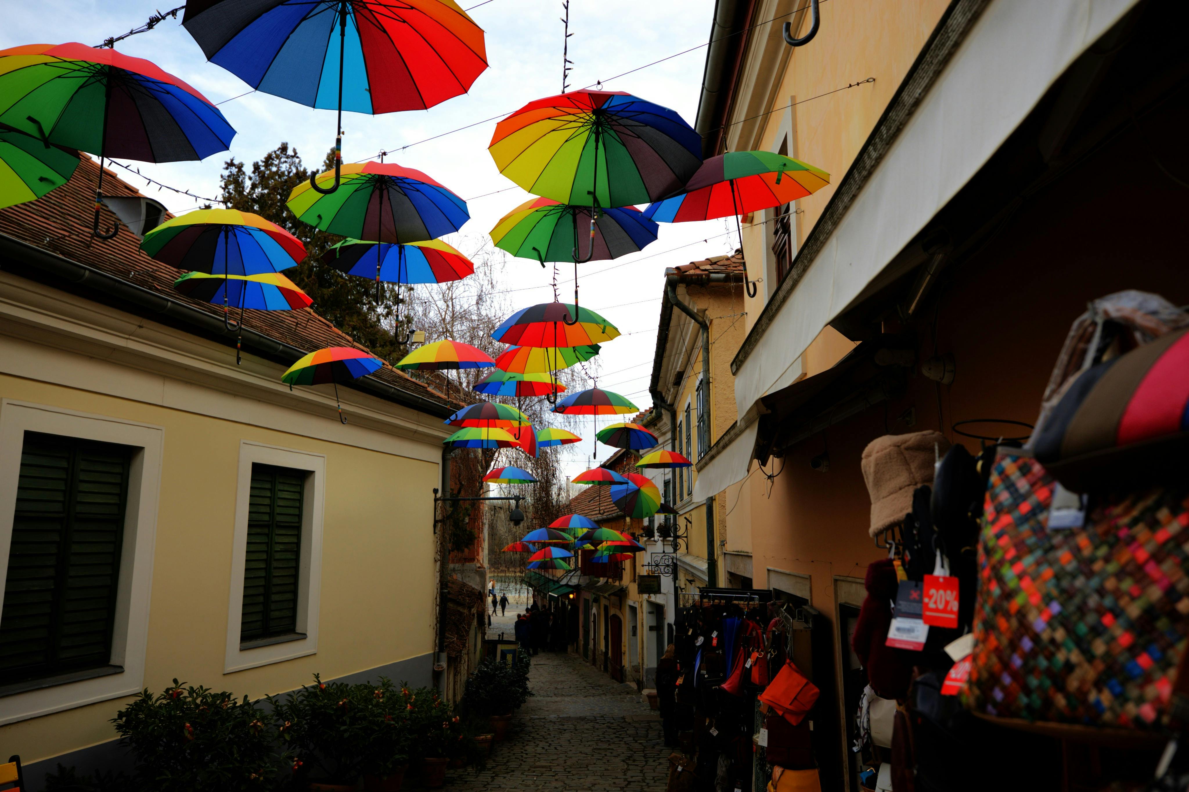 Una calle estrecha con paraguas de colores colgando, flanqueada por edificios y tiendas que exhiben mercancías.