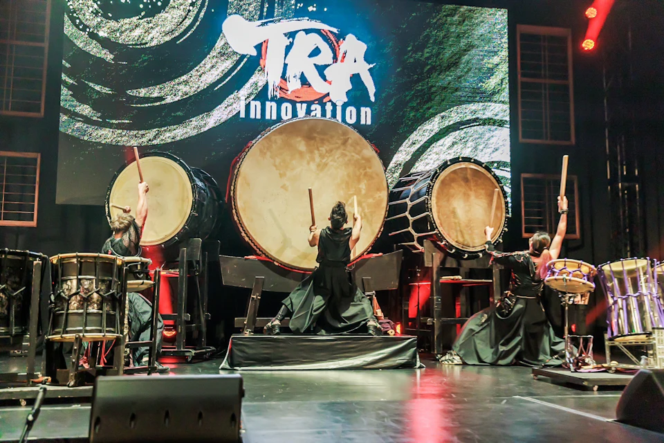A group of drummers performing on stage with large traditional drums, with colorful lighting and a backdrop displaying 'TRA Innovation.'