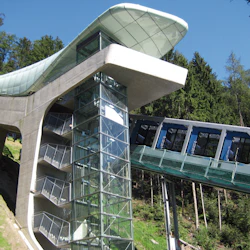 Modern linbanestation med en inglasad hiss och anslutna vagnar uppför en skogbevuxen sluttning.