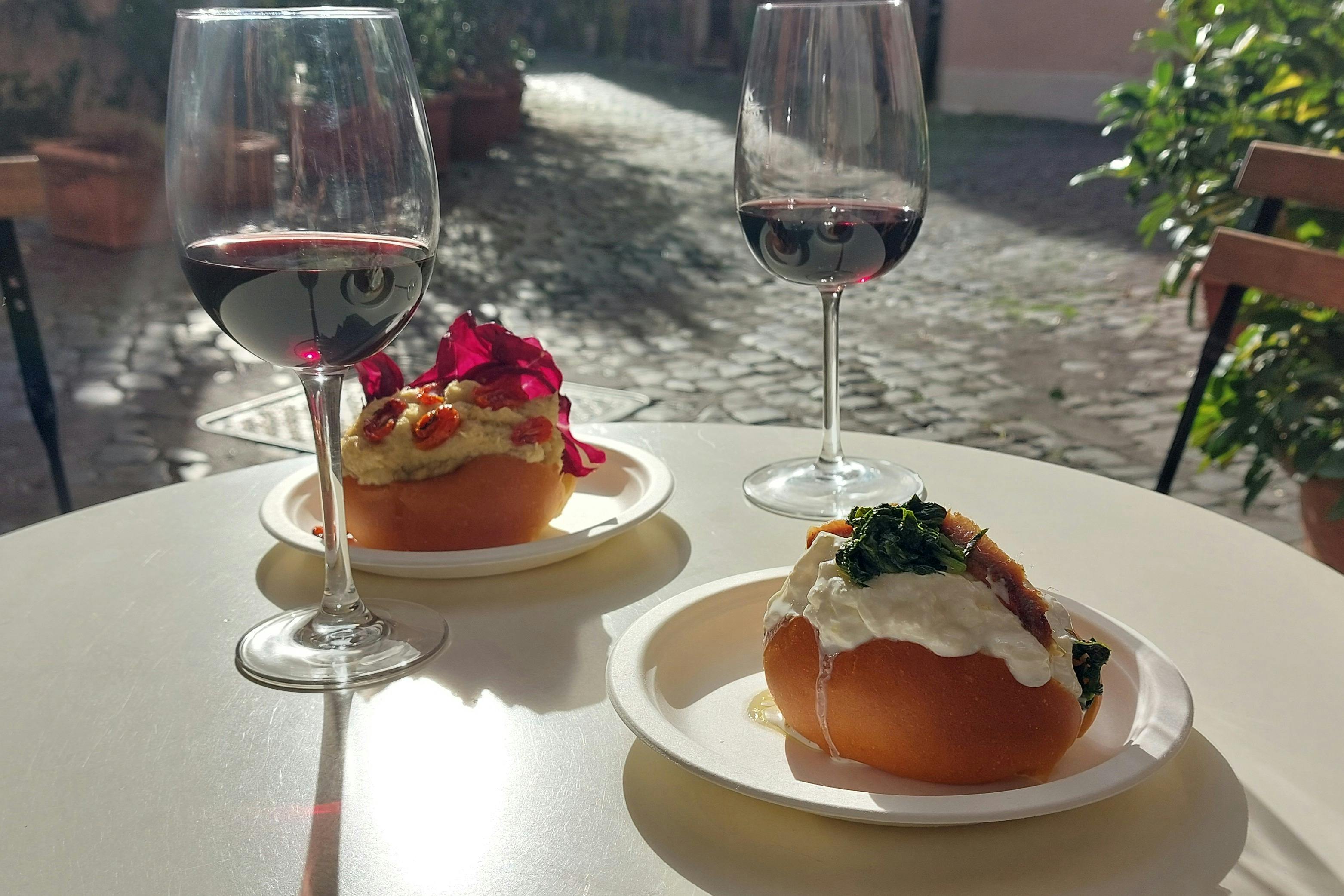 Two glasses of red wine with two plated appetizers on a white table, set outdoors on a cobblestone street.