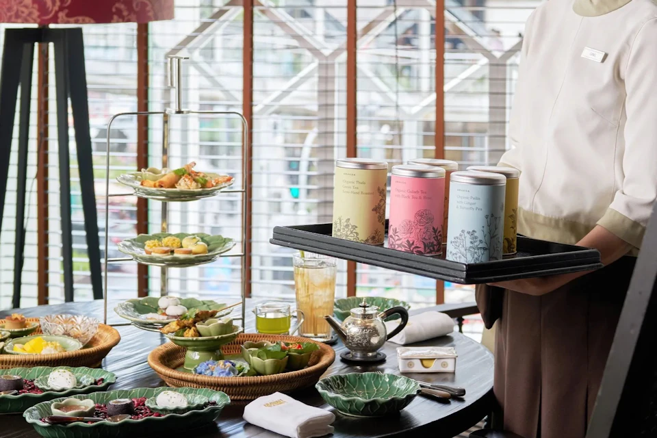 A table set with afternoon tea items, including a tiered tray, teapot, dishes, and a server holding a tray of tea tins.