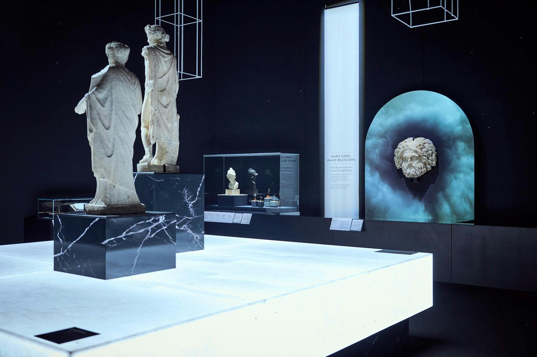 Exposition muséale présentant des statues anciennes, une vitrine contenant des artefacts et une tête sculptée sur fond bleu.