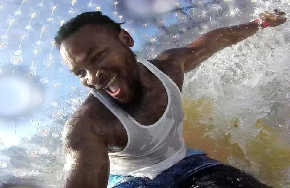 A man in a wet white tank top smiling and enjoying himself inside a transparent inflatable water ball.