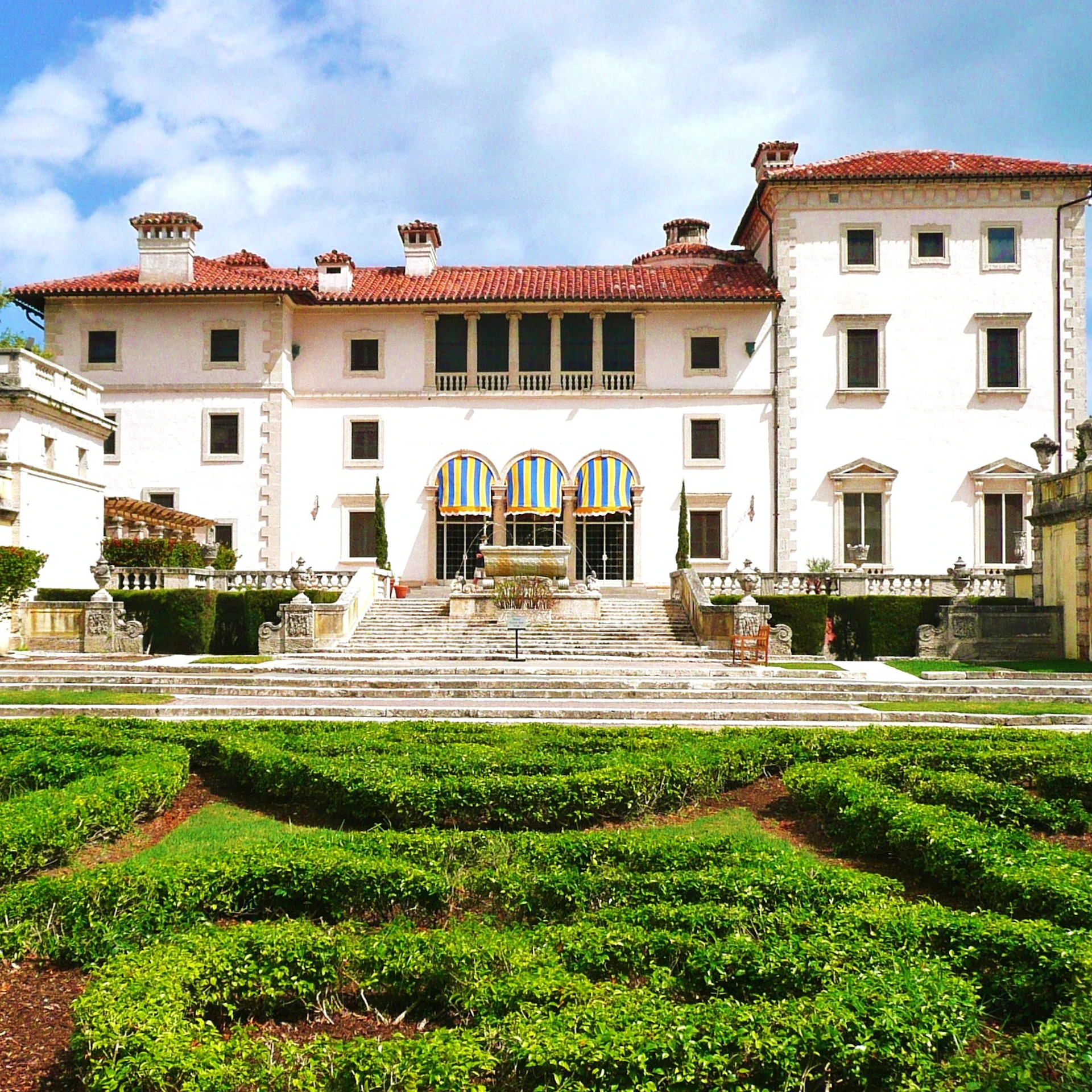 A grand white mansion with a red-tiled roof, arched windows, and manicured garden in the foreground under a partly cloudy sky.